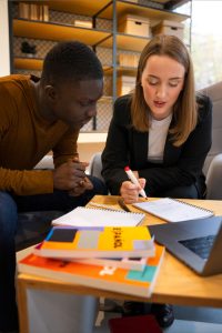 Two people sit at a table, focused on notes and a laptop. One writes in a notebook while the other leans in attentively, looking at notebooks spread out on the table.