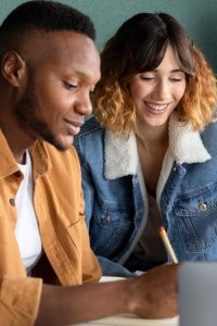 A man and a woman sit together, smiling and working on something. The man wears a brown jacket, and the woman wears a denim jacket with a shearling collar. He holds a pencil and looks at a notebook or paper.