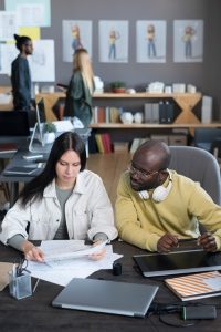Two colleagues sit at a desk in an office, reviewing papers together. The woman holds documents, while the man with glasses and headphones looks on. Other people work in the background near shelves and wall charts.